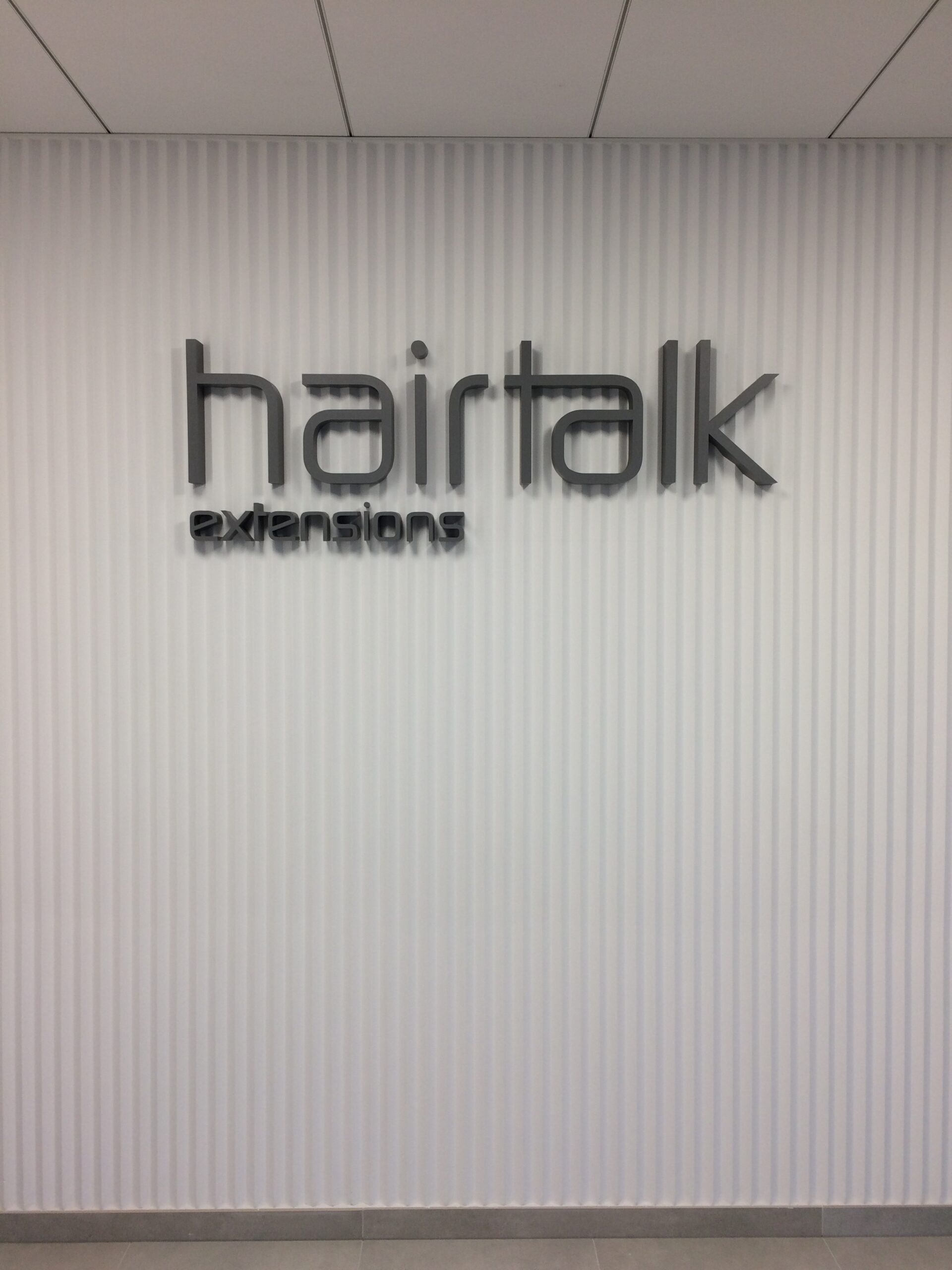 logotyp hairtalk extensions - szare litery blokowe duże i małe montowane na lamelkowej ścianie z trójkątnych wystających do przodu pionowych pasów.