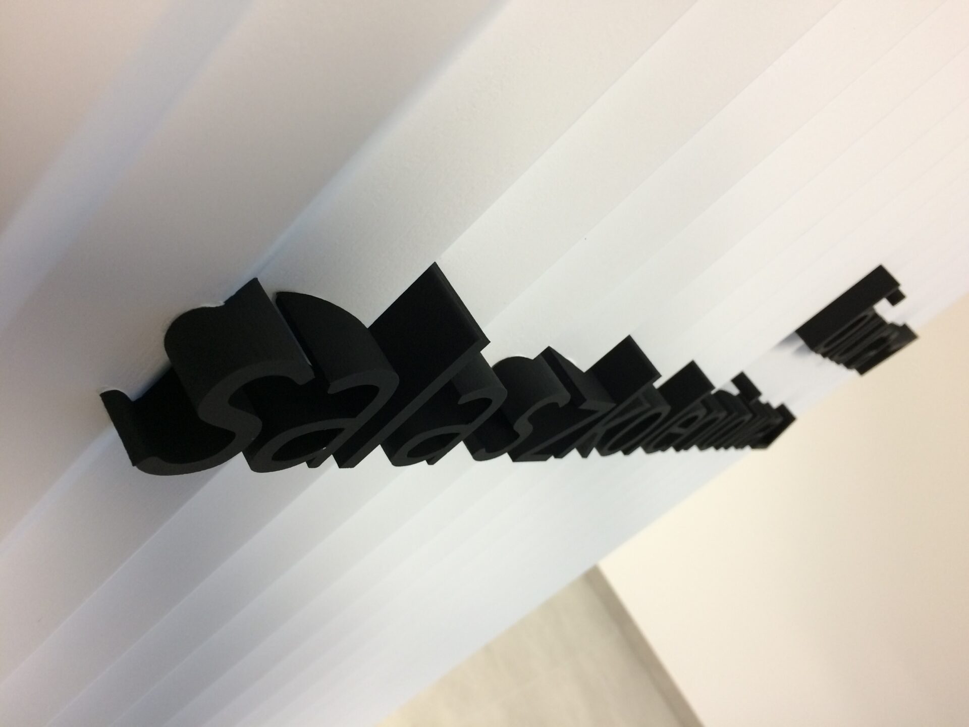 czarne dosyć małe litery blokowe wraz z odpowiednim piktogramem strzałki wskazującej kierunek, na białej ścianie lamelkowej z wystających do przodu trójkątnych pasów. biura sala szkoleniowa