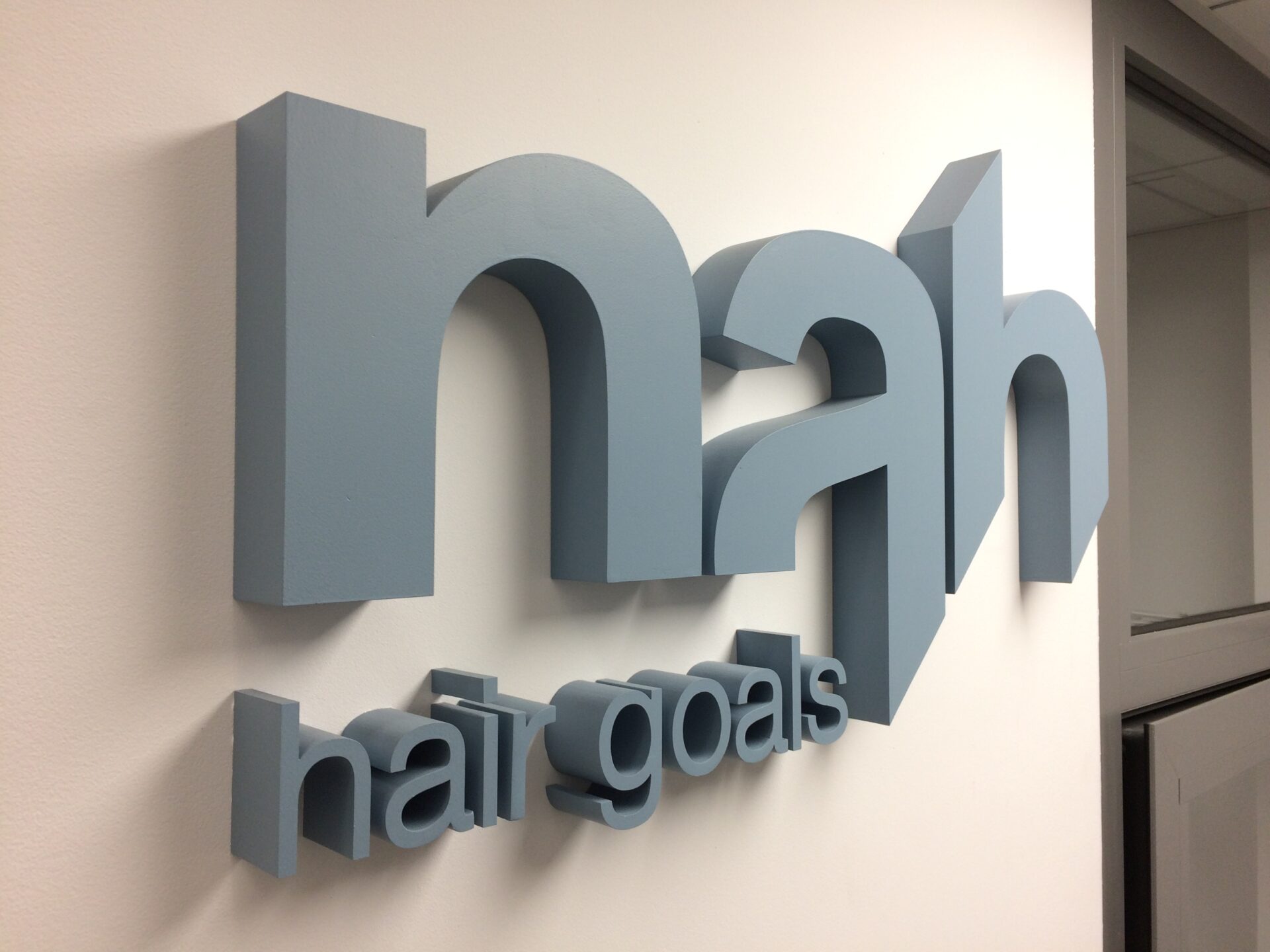 Szarozielone litery blokowe na białej ścianie: nah - duży napis w formie graficznej z powiększonym a, opdicętym 1/4 wysokości hair goals - mały napis